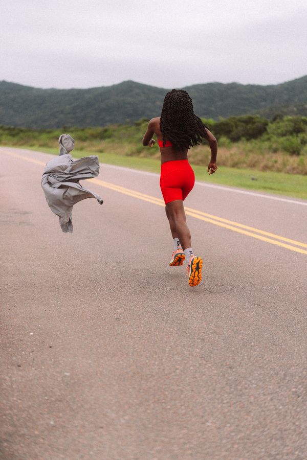 Corredora de top e shorts vermelhos correndo na estrada, com o corta-vento cinza voando